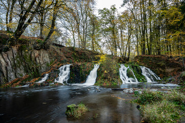 Long exposure captures a waterfall flowing smoothly through an autumn forest near Hirson, France, surrounded by colorful fall foliage.