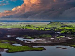 Aerial view of Iceland's landscape featuring moss covered hills, volcanic terrain, crater formations, small lakes, and a prominent hill in the midground.