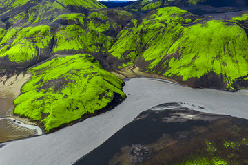 Aerial view of green moss covered hills, dark volcanic soil, and a winding grayish river cutting through Iceland's dramatic natural landscape.