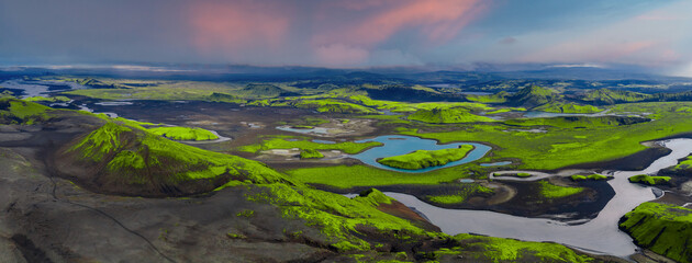 Aerial view of Iceland's landscape with green moss covered hills, volcanic craters, winding rivers, small lakes, and a cloudy sky overhead.