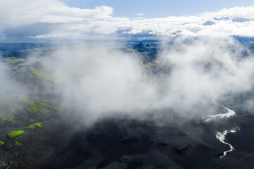 Aerial view of a winding white river cutting through dark volcanic terrain, with green vegetation, a distinct mountain, and scattered clouds in Iceland.