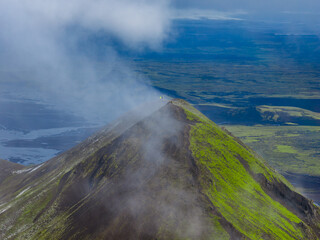 Aerial view of a green moss covered volcanic peak in Iceland, surrounded by mist, expansive plains, water bodies, and rugged distant terrain.
