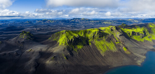 Aerial view of Iceland's volcanic terrain with a crater, steep slopes, a blue lake, green moss covered hills, and distant mountains under a cloudy sky.