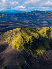Aerial view of Iceland's volcanic landscape with a moss covered ridge, dark barren terrain, distant hills, and mountains under a partly cloudy sky.