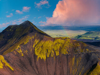 Aerial view of a volcanic mountain in Iceland with moss covered slopes, dark rock, a faint rainbow, flatlands, and distant hills under a cloudy sky.