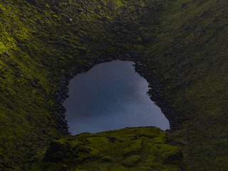 A volcanic crater in Iceland, partially filled with still water, surrounded by green moss and dark...