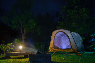 Camping tent and gas lantern on the grass next to a wood stove in the forest.