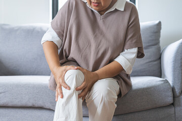 Body arthritis muscles stiff problem, aged woman suffering on leg, pain knee from disease chronic, hand massage rubbing joint injury, mature hurt or sore sitting on sofa at home. Health care people.