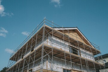 Vertical low-angle shot of tall steel scaffolding encasing a newly built wooden home with white insulation boards beneath a clear blue sky