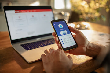Young man using a banking app on his smart phone while working from home on his laptop in the kitchen