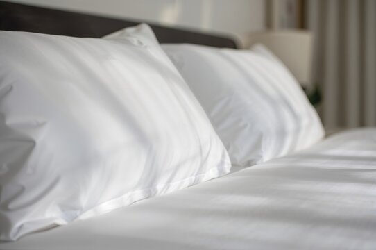 Close-up of a neatly arranged bed featuring spotless white pillows and linens, illuminated by sunlight with lens flare.