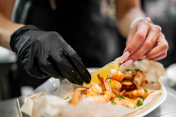 Close-up of gloved and manicured hands plating cooked shrimp with herbs and lemon on parchment paper, emphasizing food styling, hygiene, and professional kitchen preparation.