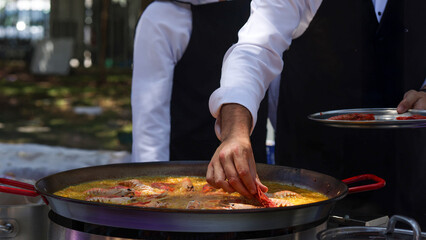 Chef preparing paella outdoors, adding langoustine to large pan