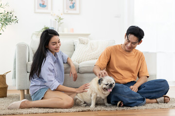 Happy Asian couple playing with adorable pet dog in cozy living room. Concept of love, relationship bonding, family lifestyle, pet companionship and joyful moments at modern home.