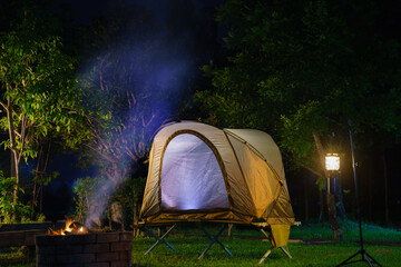 Camping tent and gas lantern on the grass next to a wood stove in the forest.