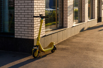 A yellow electric scooter is parked on the asphalt near the brick wall of a modern building.