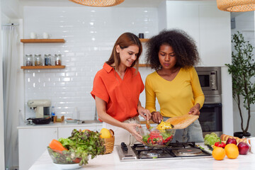 Happy LGBT lesbian couple are cooking together in a kitchen. Straight-haired women wear orange...