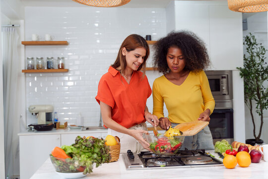Happy LGBT lesbian couple are cooking together in a kitchen. Straight-haired women wear orange shirts and curly-haired women wear yellow shirts.