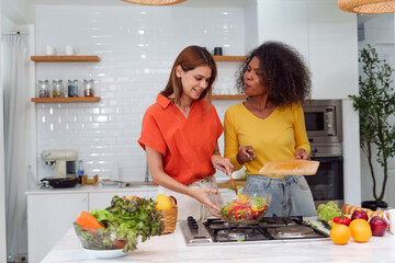 Happy LGBT lesbian couple are cooking together in a kitchen. Straight-haired women wear orange...