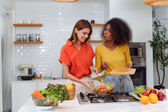 Happy LGBT lesbian couple are cooking together in a kitchen. Straight-haired women wear orange shirts and curly-haired women wear yellow shirts. They are preparing a salad