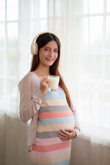 A pregnant woman is holding a glass of milk while listening to music. She is smiling, and she is...
