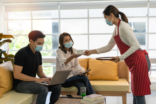 A man in black polo shirt and a woman in white shirt sitting on a couch in the coffee shop. A woman in a red apron serves a cup of coffee to a woman in a white shirt. A man using a laptop. - Powered by Adobe