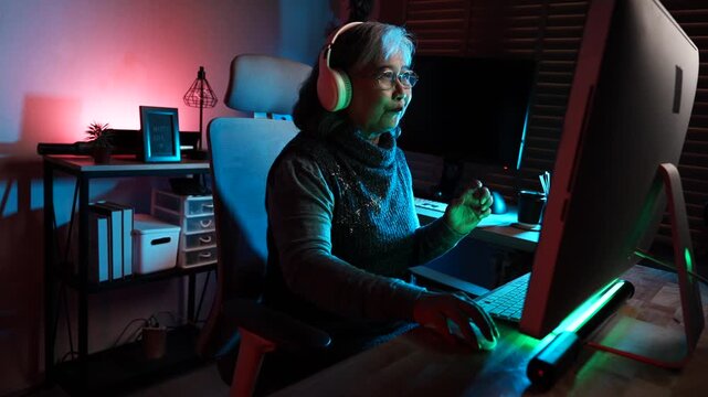 Elderly people have a hobby of playing esports. Concept of an Asian elderly woman wearing headphones happily playing online games, looking at the screen, excitedly going to a game room