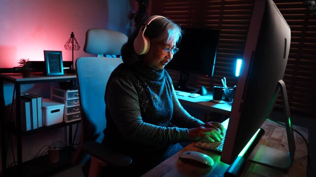 Elderly people have a hobby of playing esports. Concept of an Asian elderly woman wearing headphones happily playing online games, looking at the screen, excitedly going to a game room