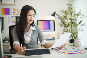 Female graphic designer reviewing printed design samples while working at computer in modern office.