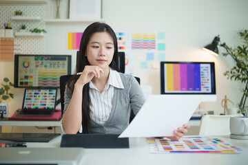 Female graphic designer reviewing printed design samples while working at computer in modern office.