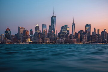 Naklejka premium Wide-angle shot of urban skyscrapers at dusk with glowing landmarks and soft blue waters in front