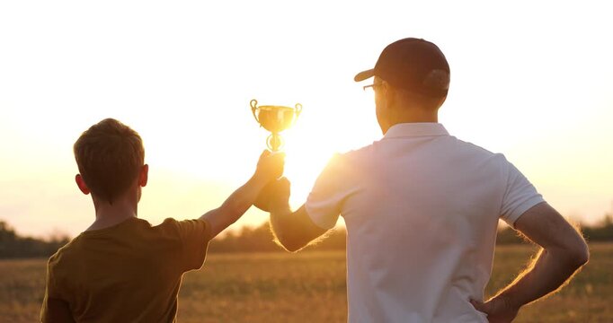 Father and child with trophy in sunset park. Father supports son proudly, parenting bond and family success. Golden light highlights joy, motivation and achievement. Father and child celebrate victory