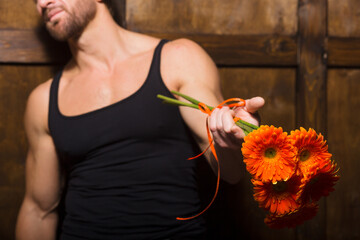 In a rustic environment, a muscular man clad in a fitted tank top gently holds a bouquet of bright orange flowers.