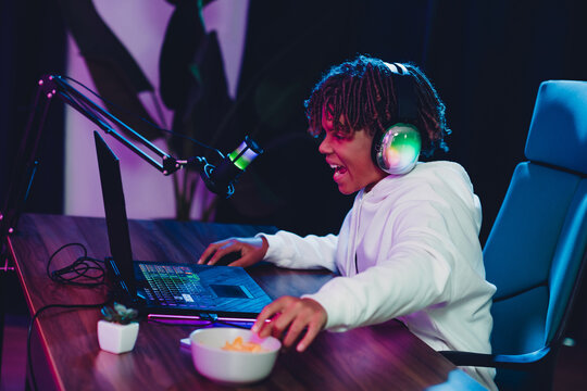 Teen gamer and content creator with headphones focuses on laptop while streaming online in an illuminated room