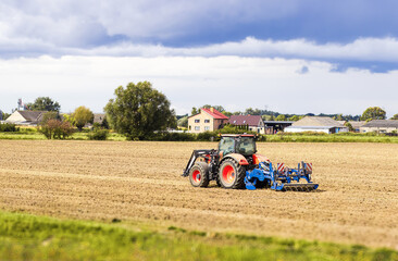 Obraz premium Tractor plowing a vast agricultural field under a dramatic sky, with distant farmhouses and trees, showcasing the essence of rural farming and land cultivation