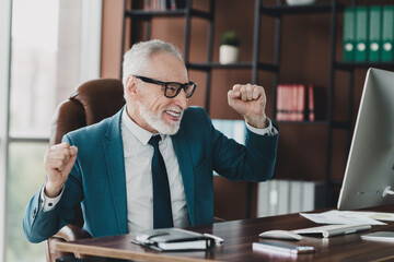 Mature businessman in formal suit celebrating success while working in a corporate office environment with a computer.