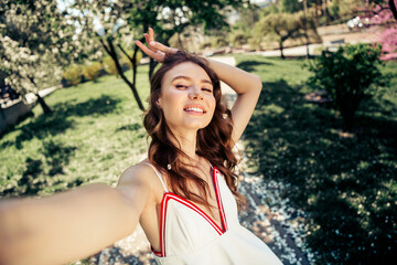 Young woman enjoying a sunny day in a blooming park, capturing a cheerful selfie while wearing a...