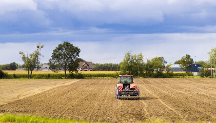 Agricultural tractor plowing a freshly tilled field under a dramatic sky, surrounded by trees and...