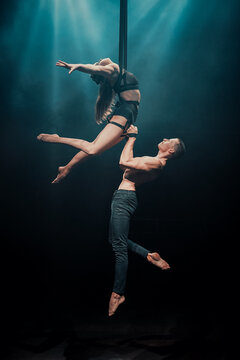Male and female aerial duo performing on straps. The man supports the woman as she arches gracefully mid-air under dramatic blue stage lighting. Horizontal image.