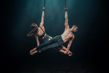 Duo aerial straps performance with male and female acrobats. Trick name: Mirrored Horizontal Hold. Both hang from straps side by side, gripping each other&rsquo;s feet under blue spotlight