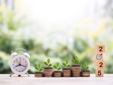 White alarm clock, Wooden blocks with the number 2025 and plants growing up on stack of coins. The concept of saving money for manage time to success business in year 2025