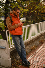 Fototapeta premium Casual man in orange vest leaning on railing and using smartphone in autumn Belgrade park