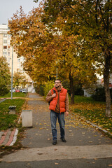 Obraz premium Man in orange vest walking along a tree-lined path covered with autumn leaves