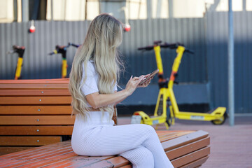 Slim girl with blonde hair using smartphone sitting on city street bench