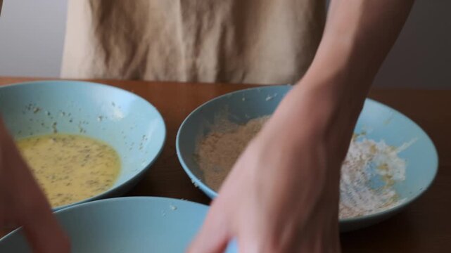 Closeup of an adult woman pressing a chicken cutlet into breadcrumbs as part of a breading station on a kitchen counter.