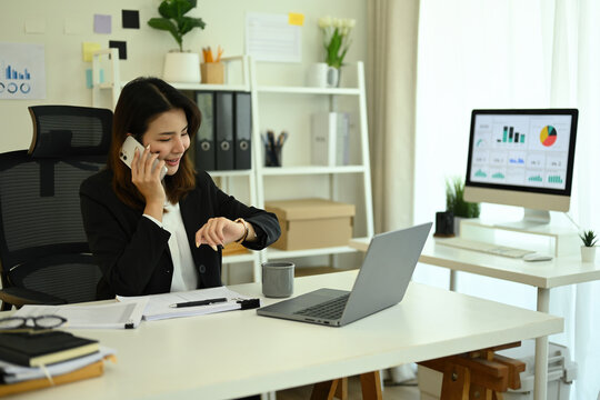 An Asian Businesswoman on phone call while checking her watch, multitasking urgent work and appointments at modern office desk