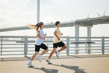Couple jogging along waterfront promenade under a modern bridge on a sunny day