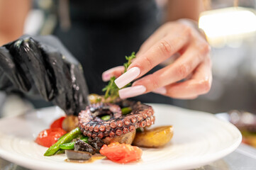 Chef plating grilled octopus with roasted vegetables and herbs. Close-up of hands in gloves and manicured nails arranging gourmet seafood dish in professional kitchen