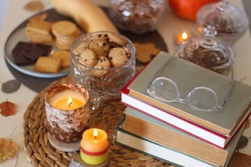 Lit candles, cookies, chocolate, nuts, tea, wine, pumpkins, books, reading glasses and autumn leaves on the table. Autumnal hygge at home. Selective focus.