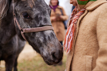 Caucasian child interacting with pony outdoors, smiling and standing close to animal, wearing warm clothing and scarf, showing gentle connection between child and horse
